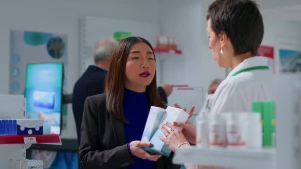 药店里的购物者手里拿着产品 讨论哪一种最适合她的需要 向专家征求意见 药剂师帮助亚洲妇女做防晒霜的推荐 — 图库视频影像