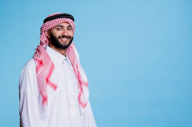 Geleneksel Arap kıyafetleri giymiş gamsız yüzlü neşeli Müslüman adam kameraya bakıyor. Gülümseyen kişi, kareli kırmızı beyaz başlıklı stüdyo resmi takıyor.