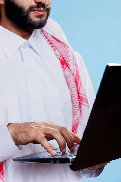 男人穿着传统的穆斯林服装在笔记本电脑键盘上打字特写 阿拉伯人站在工作室近视的便携式计算机旁 一边手写着在线信息 — 图库照片