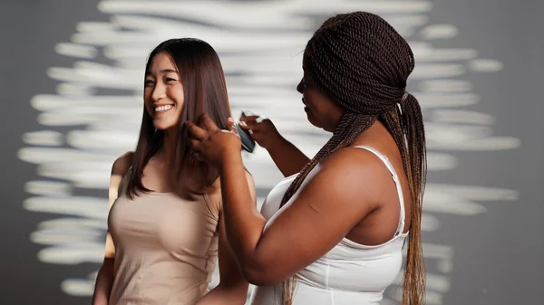 African american girl brushing hair of asian friend on camera, posing to promote friendship and wellness. Interracial women using cosmetics, advertising skincare products for ad campaign.