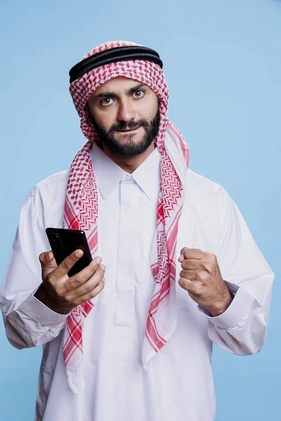 兴奋的穆斯林男子穿着传统的衣服 用智能手机 用紧握着的拳头肖像画出了不错的手势 穿着正装 手持手机 摆出优胜架势的阿拉伯人 — 图库照片