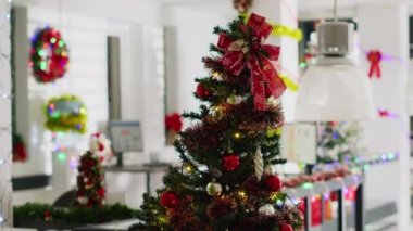 Boş ofisteki kırmızı süslemelerle süslenmiş Noel ağacı. Kış tatili boyunca Xmas dekore edilmiş iş yerlerindeki masalardaki bilgisayar monitörlerinin bulanık arkaplanı.