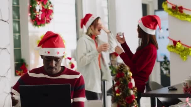 アフリカ系アメリカ人男性がクリスマスに飾られたオフィスで書類を充填し スタッフが背景に騒がしいことに気を取られました 同僚が大声で話すことにイライラする労働者 — ストック動画