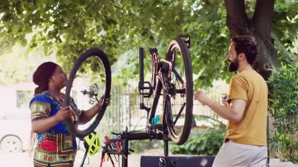若い熱狂的な白人男性とアフリカ系アメリカ人女性は 屋外の夏の趣味として自転車のホイールでメンテナンスを行います 専門装置が付いている自転車タイヤを留める多民族のカップル — ストック動画