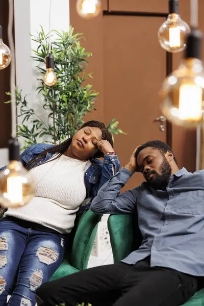 Young African American couple resting in hotel lobby after long flight, fall asleep, travelers feeling tired and exhausted, sleeping while waiting for check-in. Jet lag disorder and air travel