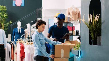 İşçi kuryeyle paketlere bakıyor, nakliye detaylarını modern butikte tartışıyor. Afro-Amerikan kurye, lojistik raporunu alışveriş merkezindeki çalışana açıklıyor..