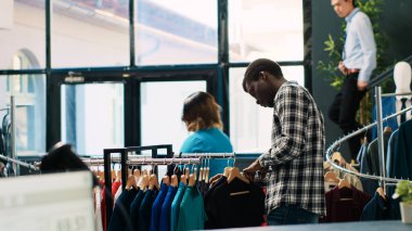 Siyah gömlek için alışveriş yapan Afrikalı Amerikalı müşteriler modern butikte moda kıyafet malzemelerini analiz ediyorlar. Alışverişkolik adam günlük giysiler alıyor, yeni moda koleksiyonu olan askılara bakıyor.