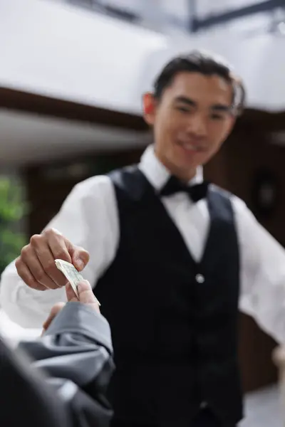 Photo showcasing happy asian bellboy receiving cash tip from tourist after assisting with luggage in hotel lobby. Guest checks in receives friendly service and gives money tip to concierge.
