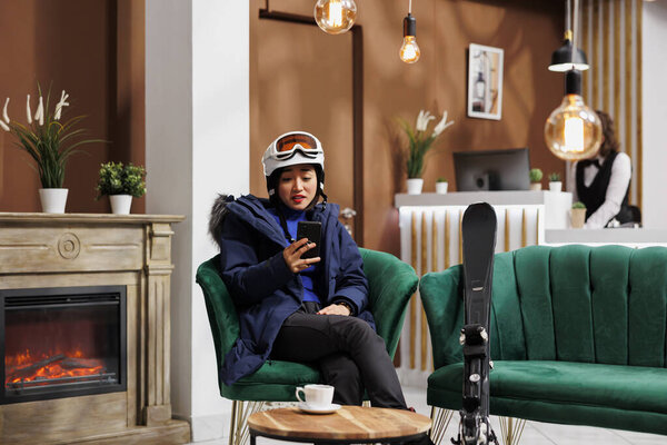 Youthful asian woman in cozy hotel lobby during her winter holiday using digital phone. Tourist wearing winter jacket ski goggles and helmet sitting on sofa with mobile device and skiing skis.