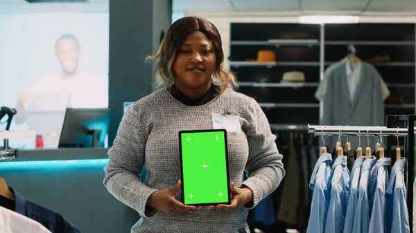 Young store manager showing tablet with green screen, advertising fashion boutique with chroma key display. Female employee using isolated mockup template with blank copyspace.