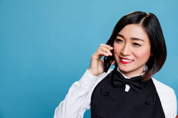 酒店笑容满面的亚洲女人接待员身穿制服 在智能手机上说话 愉快的女服务员接听电话 在手机上与餐饮服务客户聊天 — 图库照片