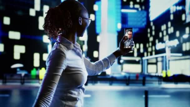 非洲裔美国妇女在晚上与男朋友一起在城市街道上漫步的视频中 在灯光昏暗的市中心用智能手机向朋友展示城市周围环境的人 — 图库视频影像