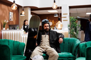 Kış sporu kıyafeti giymiş genç beyaz adam özel otelin dinlenme odasında rahat bir şekilde kayıt için bekliyor. Tatil köyü resepsiyonunda konaklamayı bekleyen aktif erkek turist.