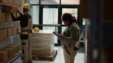 Depoda elinde tablet bulunan neşeli takım liderinin portresi, ambalajı kontrol etmek, pürüzsüz işlemleri sağlarken arka plandaki asistan karton kutulardaki paketlenmiş malları nakledilmeye hazır hale getiriyor.