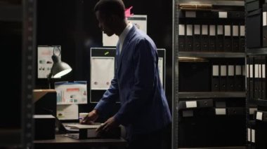 Odaklanmış Afro-Amerikan ajanı titizlikle kanıtları inceler, kayıtları araştırır ve raflardan ve laptoptan bilgi toplar. Siyahi polis ayakta duruyor ve ofis masasındaki dosyaları inceliyor..