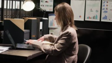 Odaklanmış beyaz kadın kanun adamı suç araştırma bürosundaki kablosuz bilgisayarla kanıtları inceliyor. Dizüstü bilgisayar, dosyalar ve özenli araştırmalar ipucu arayışına katkıda bulunur.