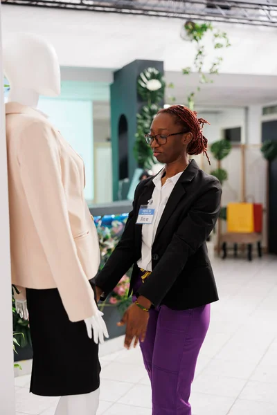 Clothing store stylist dressing mannequin in formal wear from new collection. Shopping mall fashion department african american woman assistant dressing dummy model in female jacket