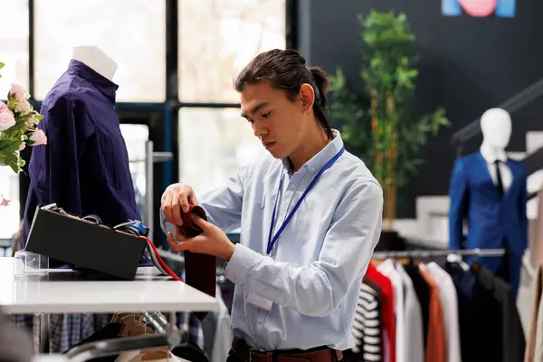 Asian customer arranging stylish accessories in modern boutique, putting belt on store shelf. Clothing store employee working with fashionable clothes in shopping mall. Fashion concept