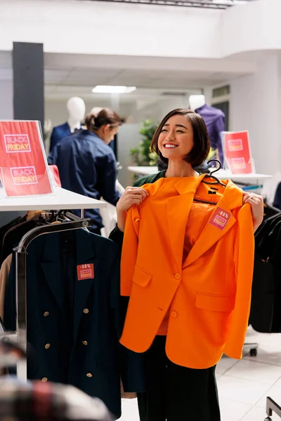 Excited happy young Asian woman holding clothing item on hanger in hands and smiling happily. Female customer feeling satisfied with low prices while shopping on black friday in fashion mall