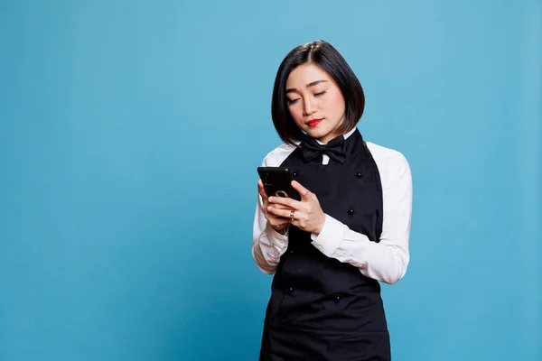 Restaurant waitress wearing uniform using smartphone to manage customer request. Young asian woman receptionist texting and reading message on mobile phone while posing in studio