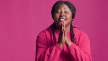 Çarpıcı siyahi kadın, ellerini birbirine uzatıp yalvarırken kameraya bakar. Pembe arka plan önünde çekici ve vücut dili talep eden genç Afrikalı Amerikalı kadın..