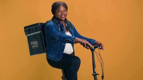 アクティブなアフリカ系アメリカ人女性が背中にバッグを持って自転車に乗って食べ物を届けようとしています 食品配達バッグを持っている若い黒人女性は 顧客に注文を取っています — ストック動画