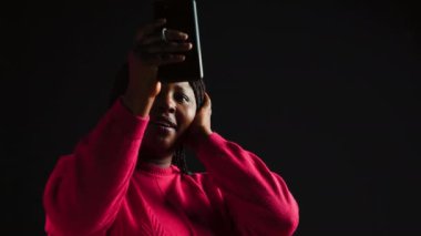 Siyah arka plandaki neşeli bir kadın akıllı telefonuyla bir video konferansında uzaktan iletişim kuruyor. Video konferans yoluyla cep telefonunda tartışma.