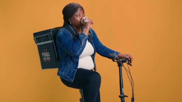 自転車の若い宅配業者は パッケージを配信しながらコーヒーを楽しむためにリラックスした瞬間を取ります 効率的な食品配送サービスを提供しながら喉の渇きを癒すために飲料を使用するアフリカ系アメリカ人女性 — ストック動画