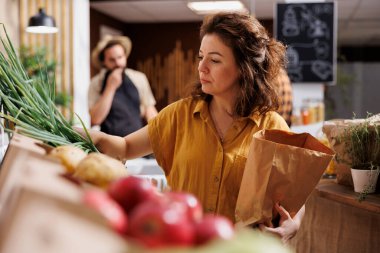 Woman in zero waste shop shopping for farm grown vegetables, picking ripe green onions. Client in plastic free local grocery shop using decomposable paper bag to avoid climate change clipart