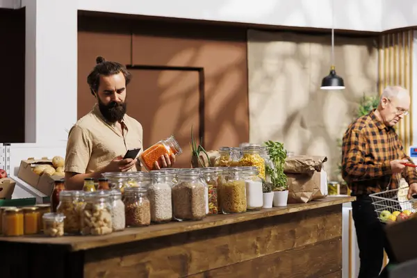 Man in zero waste store analyzing bulk products, using phone to make sure they are freshly harvested. Sustainable living customer thoroughly checking local supermarket food items are additives free