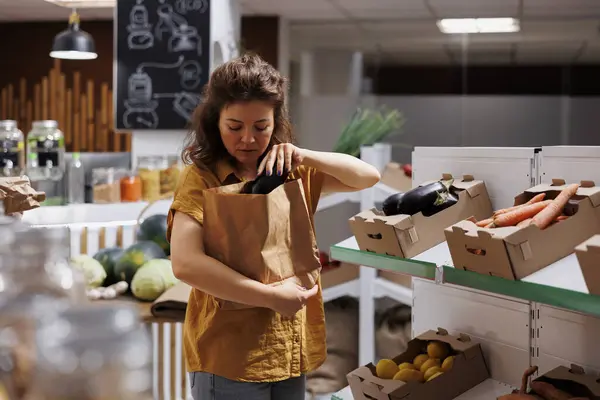 Woman in zero waste shop purchasing natural farm grown vegetables, picking ripe eggplants. Customer in plastic free local grocery shop looking to buy healthy food, using biodegradable paper bag