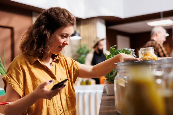 正在节食的健康女性使用智能手机 以确保零浪费超市食品适合她 本地社区生物商店的顾客确保产品是有机的 — 图库照片
