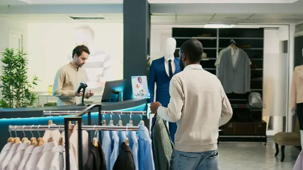 Male shopper buying formal or casual wear in boutique, standing at clothing store cash register. Young customer buying trendy merchandise and clothes, cash register commercial activity.