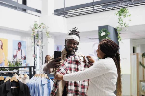 African american marketing specialists holding shirt and taking photo to post in store social media account. Fashion bloggers posing for selfie with clothes for brand promotion