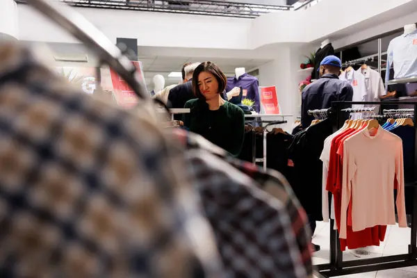 不满的亚洲女人穿着服装站在服装店里 透过衣架看衣服 女性购物者在时尚商场购物时对黑色星期五的销售感到失望 — 图库照片