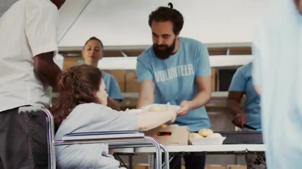 在室外食物银行 一个贫穷的白人女轮椅使用者免费获得食物和营养 身穿蓝色T恤的年轻慈善工作者向不幸的人分发新鲜水果和热饭 — 图库视频影像