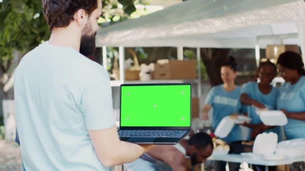 孤立した緑色のスクリーンを搭載したラップトップを運ぶ若い男性の詳細なビューは ボランティアのグループが貧しい人々を助けます 男性チャリティワーカーが保有する染色体モックアップテンプレートでデジタルデバイスに焦点を当てる — ストック動画
