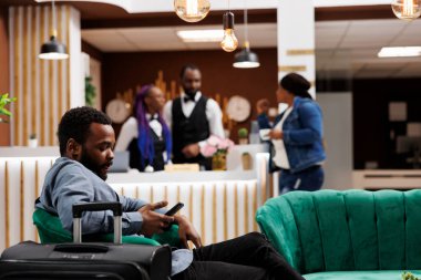 Afrikalı Amerikalı gezgin otel lobisinde oturmuş arkadaşlarıyla sohbet ederken otelin girişini bekliyor, ücretsiz kablosuz ağ kullanıyor. Bagajlı bir turist resepsiyonda bekliyor.