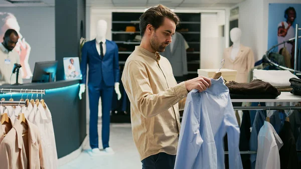 Male customer analyzing fashion collection in boutique and formal wear, young adult shopping for trendy clothes to increase wardrobe. Caucasian man looking at merchandise hanging in store.