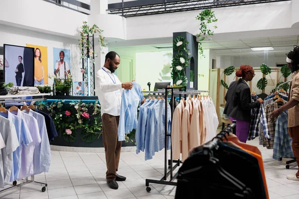 Clothes shop african american employee hanging shirt on rack. Fashion garment showroom assistant checking formal wear while working in store and choosing apparel for customer