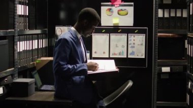 Afro-Amerikan müfettişin portresi kanıt odasında duruyor, dava dosyalarını okuyor. Loş bir ofiste, erkek müfettiş şüpheli ifadeleri inceler ve kameraya bakar. Yavaş çekim, yakınlaştır.