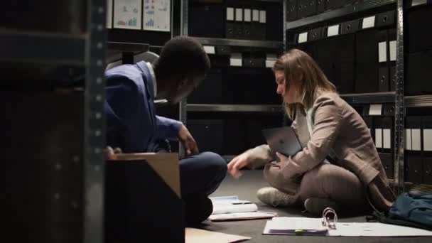 在进行犯罪调查的过程中 黑人警察和女警官带着文书工作和手提电脑坐在办公室地板上 多种族调查员审查和辩论案件证据和提示 — 图库视频影像