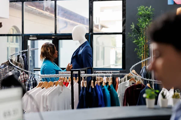 Stylish woman shopping in clothing store, looking at mannequin with formal suit. Shopaholic african american woman analyzing fashionable merchandise fabric in modern boutique. Fashion concept
