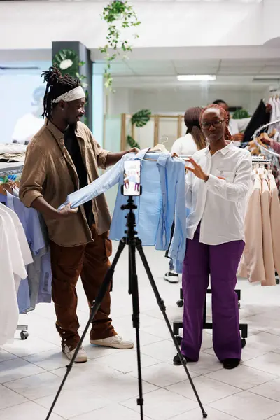 Social media influencer and clothes store assistant filming live video and showcasing shirt from latest arrival. African american blogger and shop manager promoting male formal wear on smartphone