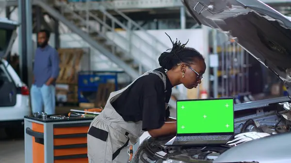 Chroma key laptop in garage workplace sitting on broken automobile. Mockup device in car service next to african american mechanics in background refurbishing damaged vehicles
