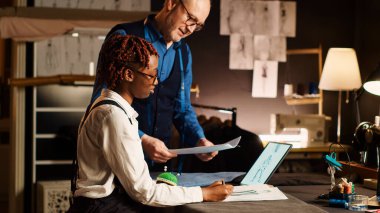 Terzi ve öğrenci atölyede eskizlere bakıyor, giyim markası yaratmak için çıraklık öğreniyor. Deneyimli tasarımcı, terzilik atölyesindeki stajyere yeteneklerini açıklıyor..