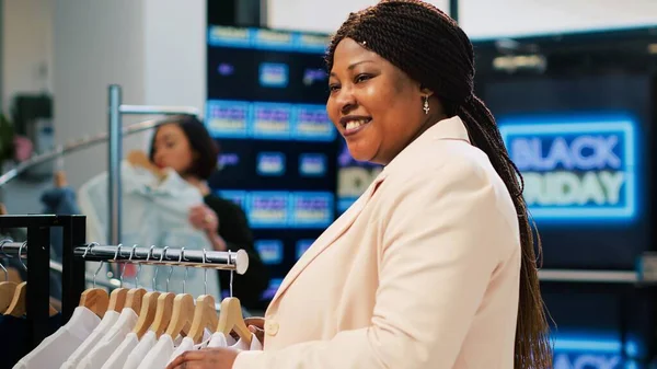 Client browsing through clothes racks on black friday, clothing store outlet with clearance section and promotional sales. African american woman shopping for discounted items.