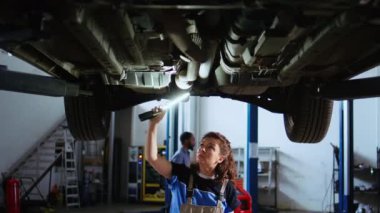 Sertifikalı teknisyen garajda askıya alınmış araba üzerinde çalışıyor. Rutin bakım sırasında bileşenleri kontrol ediyor. Otomobil tamircisi aracın altında yürüyor, iş ışığı kullanarak inceliyor.