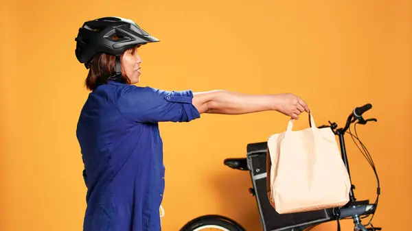 BIPOC food delivery worker reaching order address, waiting for customer to answer door. Courier woman greeting client, offering takeaway lunch bag, isolated over orange studio background