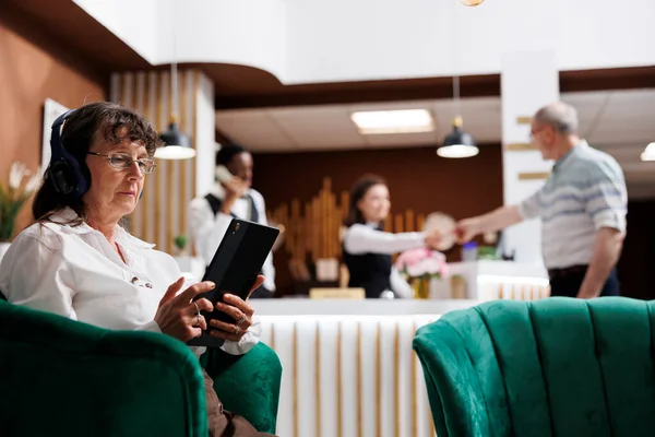 Elderly female tourist wearing wireless headset and using a digital tablet while on vacation. At hotel foyer, receptionist serves male customer with check-in. Great hospitality and technology.